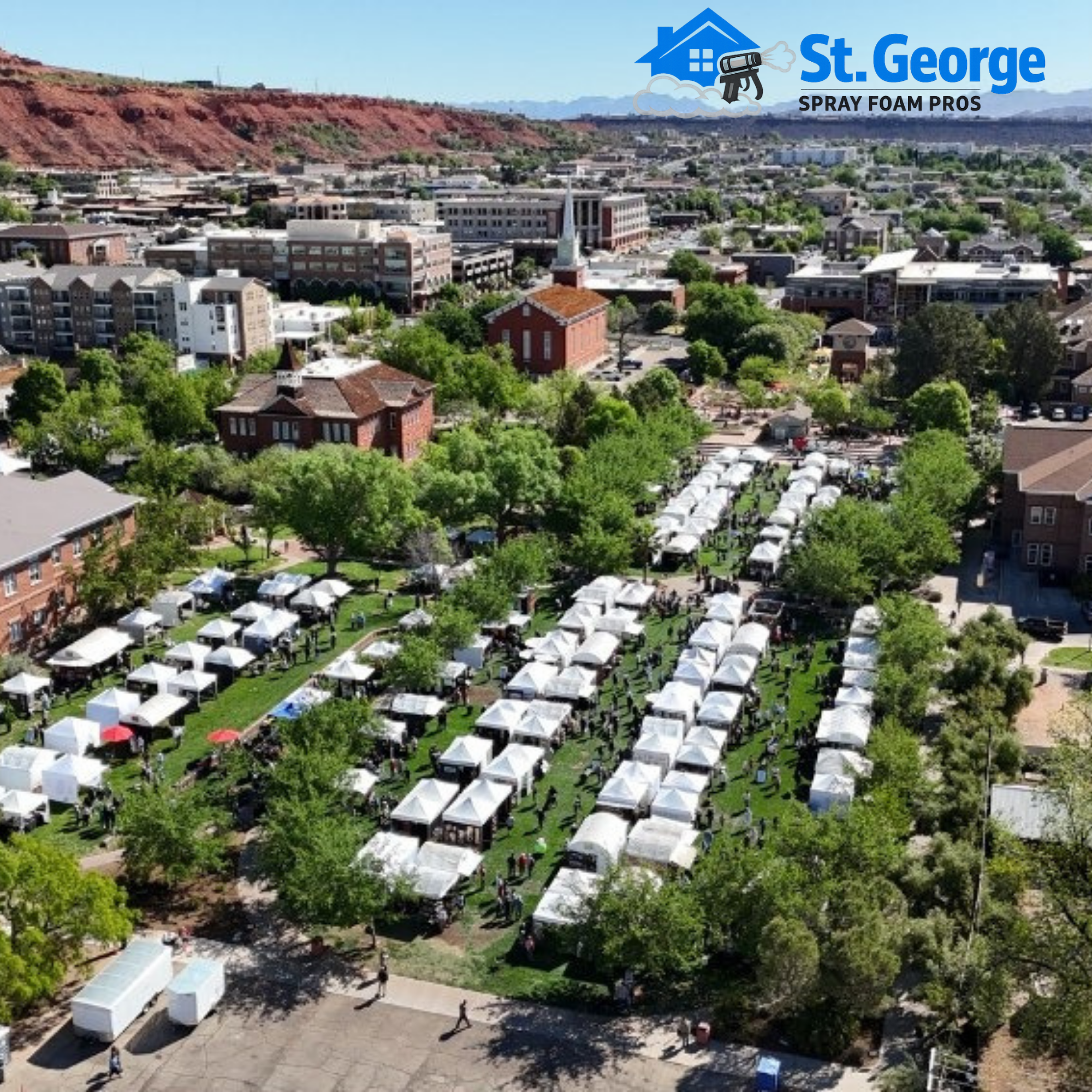 Aerial view of downtown St. George, Utah with the historic St. George Tabernacle and rows of white event tents in a park, framed by red sandstone bluffs